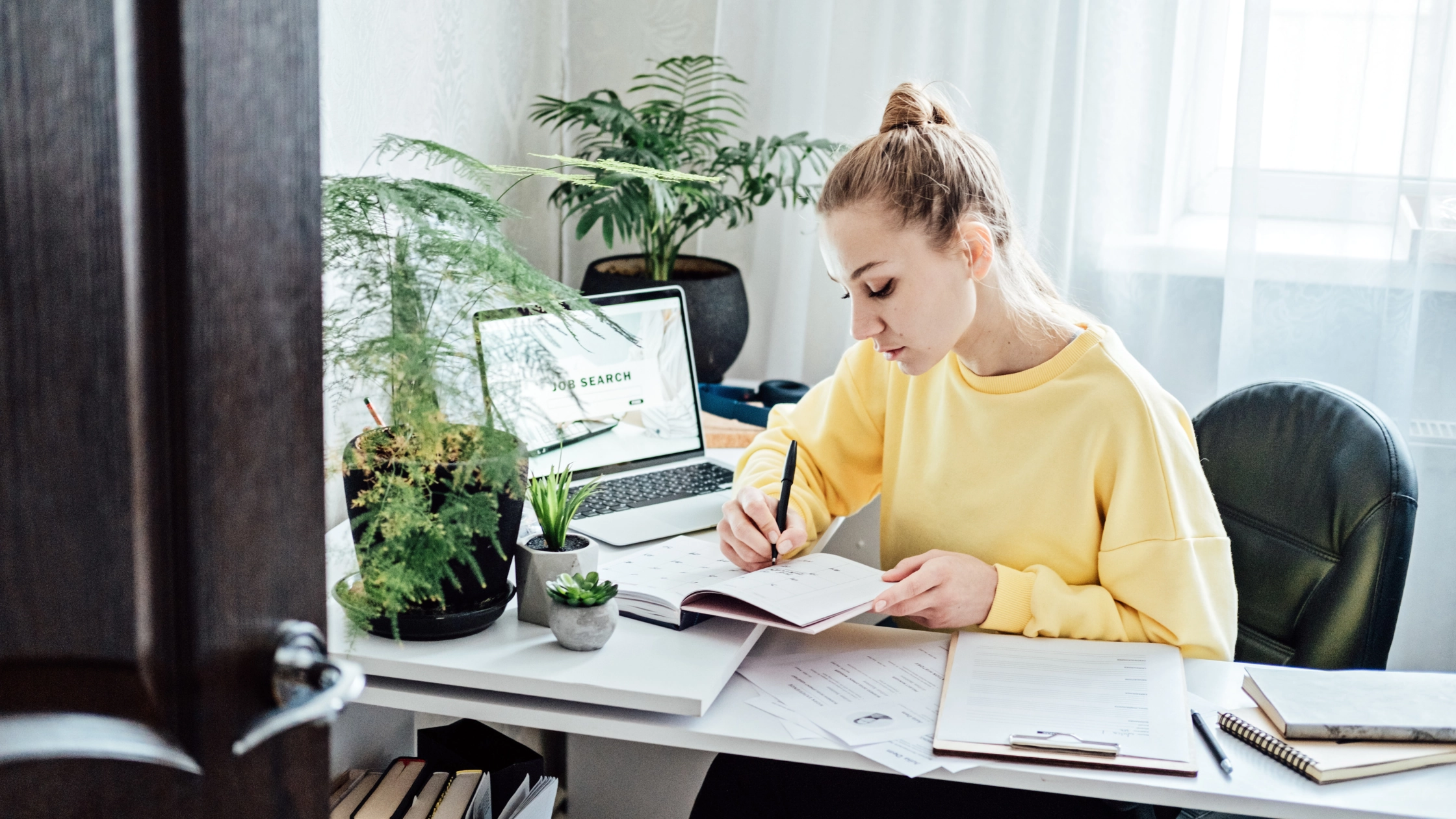 A woman at a desk with assorted documents, writing notes with a laptop displaying "Job Search" beside her.