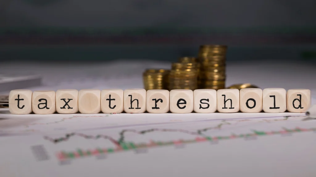 Small wooden letter blocks forming the words "tax threshold" placed on paper financial charts with gold coins in the background.